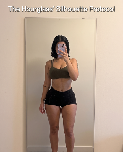 Woman in an olive sports bra and black high-waisted shorts taking a mirror selfie showing her hourglass figure workout results